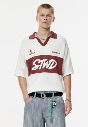 White and maroon polo with a v-neck, featuring a large white "STWD" logo and red accents. Worn with light wash jeans and accessories.