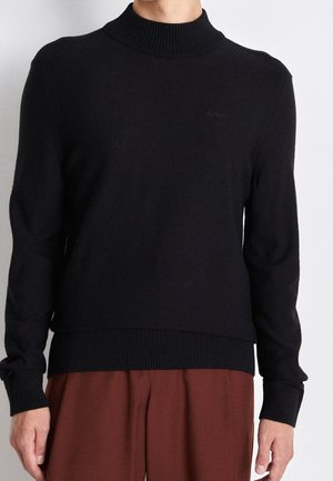 Personne portant un pull à col roulé côtelé noir avec le logo "BOSS" et un pantalon marron, debout devant un fond clair uni.