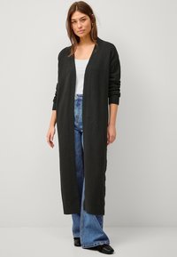 Langer schwarzer Cardigan aus einem strukturierten Stoff mit offenem Frontbereich, gerippten Bündchen und einer lockeren Passform, kombiniert mit hochgeschnittenen blauen Jeans.