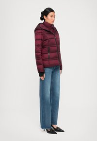 Woman wearing a burgundy puffer jacket with a hood, blue wide-leg jeans, and black pointed-toe heels, standing against a white background.