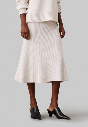 Frau trägt einen cremefarbenen, gerippten Strick-Midirock, passenden Pullover und schwarze spitze Mules mit Kegelhochabsätzen vor einem grauen Hintergrund.