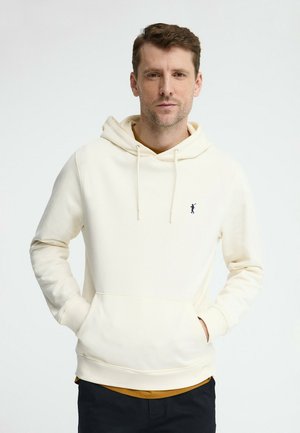 Uomo con capelli castani corti che indossa una felpa con cappuccio color crema con un piccolo logo scuro, mani nelle tasche, in piedi davanti a uno sfondo bianco semplice.