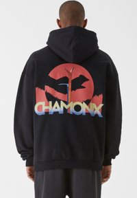 Fekete kapucnis pulóver, amelynek hátán egy nagy, színes grafika látható kör alakú mintával, hegyvonalzattal és az alsó részén "CHAMONIX" felirattal.