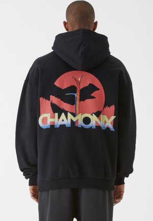 Sort hoodie med et stort farverigt grafisk design på ryggen, som har et cirkulært mønster, bjergsilhuet og teksten "CHAMONIX" nederst.