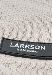 Béžová tkanina s textúrovaným vzorom, na ktorej sa nachádza čierny obdĺžnikový štítok s nápisom "LARKSON HAMBURG" v bielych písmenách.