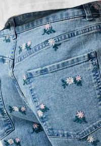 Jeans in denim azzurro chiaro con piccoli fiori ricamati rosa e bianchi sparsi sul tessuto e una tasca posteriore visibile.