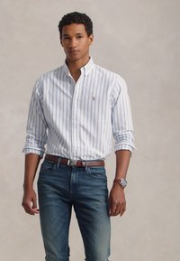 Camisa de botão com riscas verticais azuis e brancas, mangas longas, combinada com jeans escuros e um cinto castanho. Relógio casual no pulso.