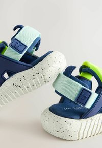 Dunkelblaue Sandalen mit verstellbaren aqua-farbenen Riemen und einem grünen Akzent. Sie verfügen über eine gesprenkelte weiße Sohle mit einer strukturierten Gummi-Oberfläche.
