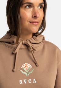 Brun hoodie med dragsko i halsen, med blommönster broderat i orange och grönt. Tyget ser mjukt och mysigt ut, lämpligt för avslappnad användning.