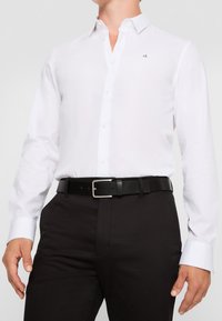 Camisa blanca de vestir con mangas largas, cuello abotonado y un logo sutil. Combinada con un cinturón negro y pantalones oscuros, exhibiendo un diseño clásico.