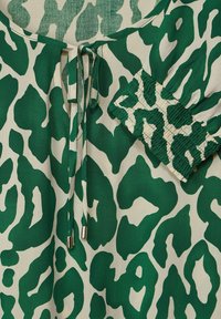 Primo piano di un tessuto animalier verde e crema con un nodo sulnesso e dettagli arricciati elasticizzati sul polsino della manica.