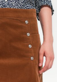 Brown corduroy skirt with silver snap buttons along the side, featuring a side pocket and ribbed texture, paired with a patterned shirt.