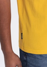 Gul bomulds t-shirt med rund kant, der har en sideetiket, vist med en delvist synlig arm mod en denimbaggrund.