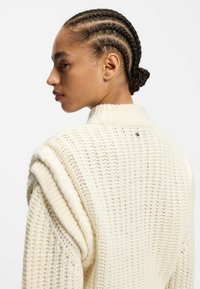 Cream-colored knitted sweater featuring a textured pattern, a high collar, and braided accents on the shoulders. Small logo detail on the back.