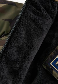 Schwarzes Fleecefutter innen in einer mit Reißverschluss versehenen Jacke, mit Tarnmuster außen und einem rechteckigen Patch mit blauer Schrift auf grünem Hintergrund.