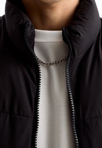 Černá prošívaná bunda s vysokým límcem, vyrobená z texturované látky a se zipem. Pod ní je bílá žebrovaná košile se stříbrným řetízkovým náhrdelníkem.