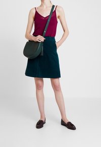 Burgundy toppi ilman hihoja, jossa on pitsireunus, tummanvihreä corduroy-hame, ruskeat loaferit ja pyöreä vihreä laukku, jossa on tupsuyksityiskohta.