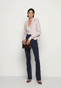 Kvinne står mot en enkel bakgrunn iført lyse rosa blazer, hvit topp, marineblå bukser, holder en mørk clutch og har på seg spisse hæler.
