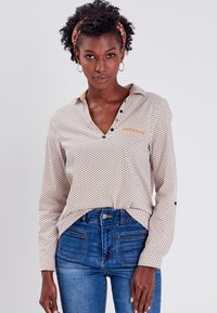 Blusa de manga larga en Beige claro, con un pequeño patrón negro y naranja, frontal abotonado y detalle de cuello en contraste. Combinada con jeans azules.