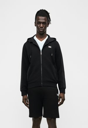 Uomo con pelle scura e dreadlocks che indossa una felpa nera con cerniera sopra una maglietta bianca a V e pantaloncini neri davanti a uno sfondo neutro.