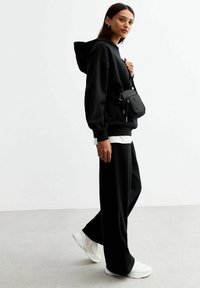 Felpa nera oversize e pantaloni larghi abbinati. Camicia bianca a strati all'orlo. Borsa a spalla nera. Scarpe sportive bianche. Sfondo minimalista.