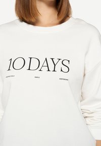 Sudadera blanca con un texto negro en negrita que dice "10 DAYS" y texto adicional más pequeño debajo, confeccionada con un tejido suave y texturizado, con un ajuste relajado.