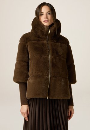 Veste en fausse fourrure marron avec un col haut, des manches courtes, un motif matelassé horizontal et une fermeture éclair à l'avant. Portée sur une jupe plissée marron.