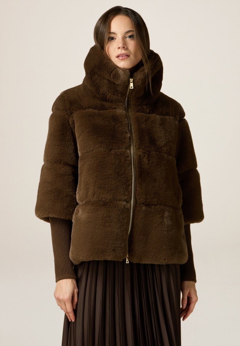 Giacca in faux fur marrone con colletto alto, maniche corte, motivo trapuntato orizzontale e zip frontale. Indossata sopra una gonna pieghettata marrone.