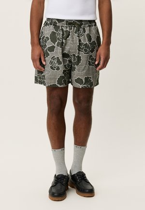 Zielone spodenki w kwiatowy wzór z elastycznym pasem i ukośnymi paskami. Noszone z szarymi skarpetkami i czarnymi butami.