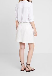 Valkoinen pellavahame, jossa on laskoksia, yhdistettynä vaaleanväriseen, napitettuun paitaan ja mustiin remmisandaaleihin, nähtynä takaa.