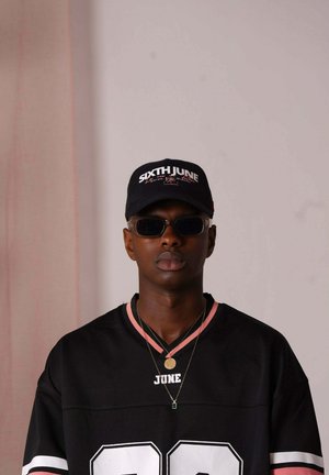 Zwarte sporttrui met roze accenten en witte "22" tekst, gecombineerd met een zwarte pet met het "SIXTH JUNE" logo en een zonnebril. Twee kettingen zichtbaar.