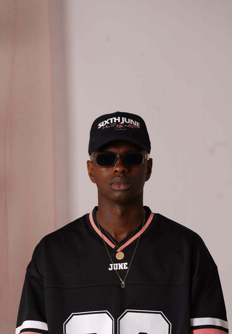 Maillot de sport noir avec des accents roses et le texte blanc "22", associé à une casquette noire avec le logo "SIXTH JUNE" et des lunettes de soleil. Deux colliers visibles.