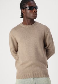Beiger Strickpullover mit rundem Halsausschnitt, gerippten Bündchen und Saum, aus einem strukturierten Stoff gefertigt. Über hellen Denim getragen.