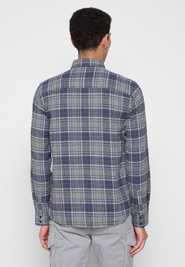 Szara flanelowa koszula w kratę z granatowymi i czerwonymi akcentami, długimi rękawami, standardowym kołnierzykiem i zaokrąglonym dołem. Tekstura materiału wydaje się miękka i fakturowana.