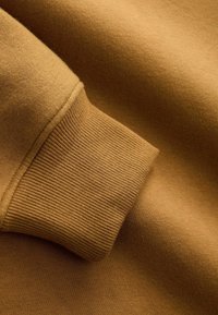 Felpa in cotone marrone caratterizzata da un design con polsini a costine e una texture liscia, mostrata da un'inquadratura ravvicinata che evidenzia i dettagli delle cuciture.
