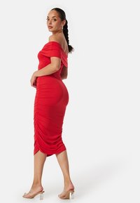 Robe rouge, décolletée, ajustée avec une texture plissée le long du corps, longueur genou. Associée à des sandales à talons transparents. Tissu lisse et extensible.