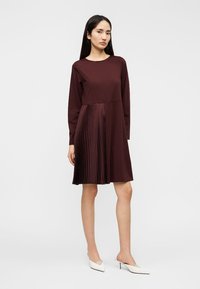 Μακρυμάνικο μπεζ φόρεμα με πλισέ κάτω μέρος, διαθέτει λείο πάνω μέρος και διακριτή μέση. Κατασκευασμένο από απαλό ύφασμα.