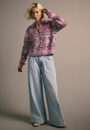 Rosa Strickjacke mit geometrischen Mustern, einem Kragen und Knöpfen, kombiniert mit weit geschnittenen hellblauen Jeans und beigen Schuhen.