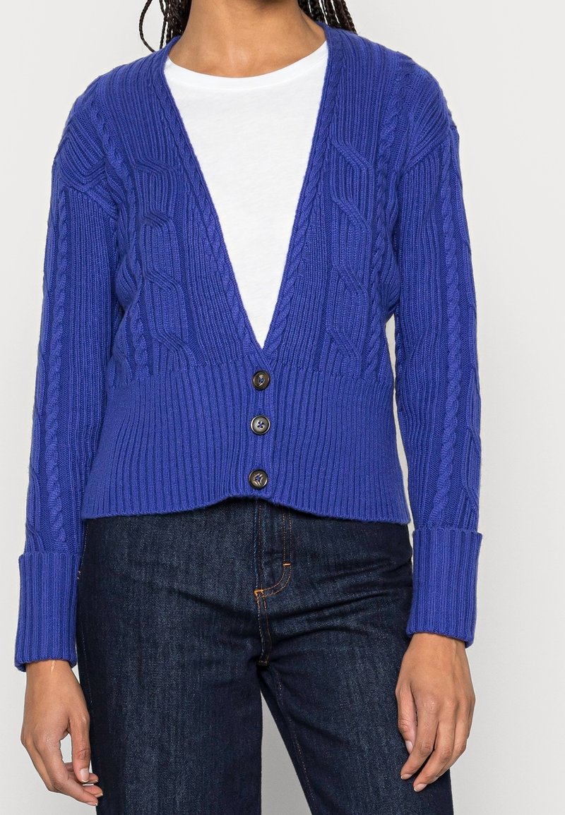 Blå stickad cardigan med djup V-ringning, ribbad nederkant och texturerad kabelstickad design, med tre knappar fram och långa ärmar.