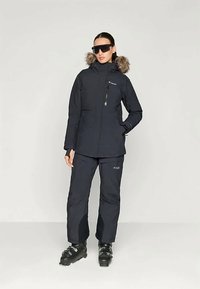 Jachetă de schi bleumarin cu glugă căptușită cu blană, buzunar cu fermoar în față și pantaloni asortați. Pur și simplu cu bocanci de schi și ochelari de soare. Material neted și izolat.