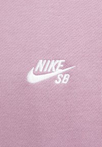 Roza pletena tkanina z belim izvezenim logotipom "Nike SB" z značilnim pasom, z gladko teksturo in enakomernim šivom.