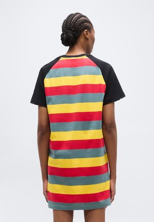 Frau mit geflochtenem Haar, die ein kurzärmeliges Kleid mit horizontalen rot-, gelb- und blau-grünen Streifen trägt, von hinten gezeigt.