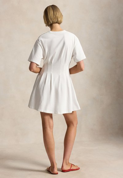 Γυναίκα που στέκεται με την πλάτη προς τα εμάς, φορώντας λευκό φόρεμα με κοντά μανίκια και κόκκινα επίπεδα σανδάλια, σε ουδέτερο φόντο.