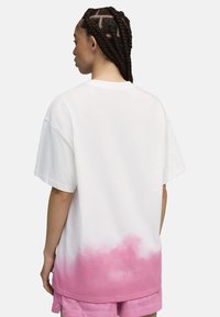 Mujer con el cabello trenzado, vistiendo una camiseta blanca de gran tamaño con un patrón de nubes rosas en la parte inferior y pantalones cortos de malla rosa, de espaldas.