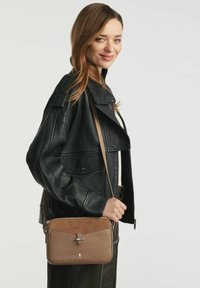Młoda kobieta w czarnej skórzanej kurtce, trzymająca brązową torbę na ramię z zapięciem, uśmiechająca się na tle jednolitego jasnego tła.
