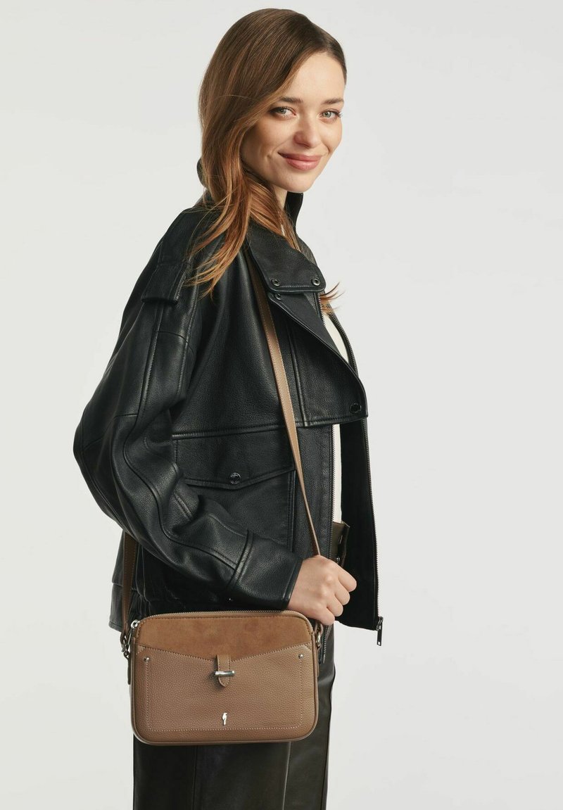 Młoda kobieta w czarnej skórzanej kurtce, trzymająca brązową torbę na ramię z zapięciem, uśmiechająca się na tle jednolitego jasnego tła.