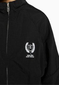 Mustat takki, jossa on korkea kaulus, hopeinen vetoketju ja brodeerattu logo, jossa on teksti: "tiimi liekki. NAUTI TIEN TÄSTÄ" sekä koristeellinen seppele.