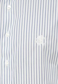 Biała koszula zapinana na guziki z niebieskimi pionowymi paskami, z małym wyszywanym logo i okrągłymi plastikowymi guzikami. Gładka faktura tkaniny.