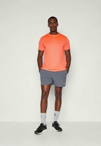 Mann som står med hendene i lommen, iført en lysoransje t-skjorte med korte ermer, grå shorts, hvite sokker og svarte joggesko mot en ensfarget bakgrunn.