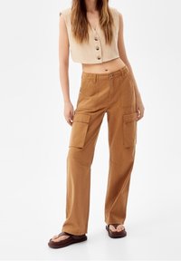 Beiga cargobyxor med flera fickor, tillsammans med en ärmlös beige topp med V-ringning och knäppning framtill. Bärs med bruna sandaler.